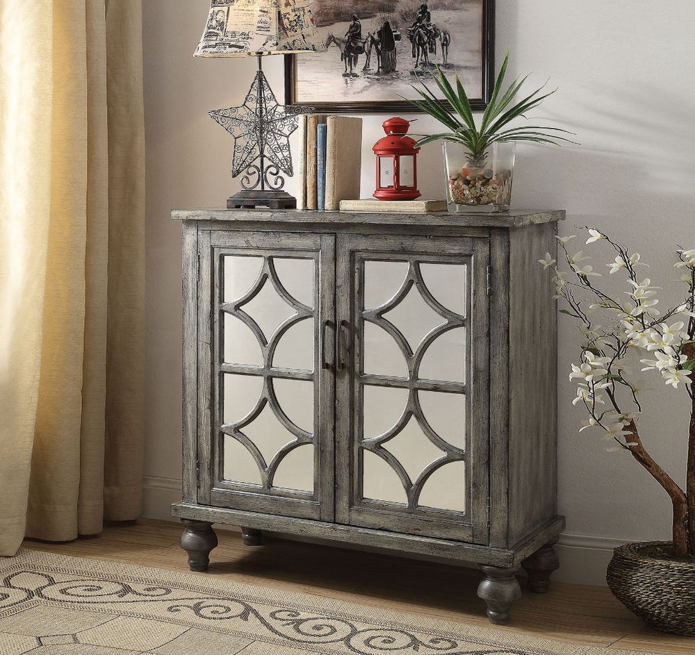 The transitional Velinka Console Table features a weather gray finish with mirrored glass doors and a diamond trim inlay. Two cabinet doors open on the rectangular wood console table to reveal ample shelf space for additional storage. The modern style cab