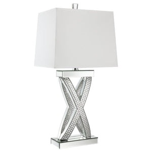 This table lamp features a sculptural X-shaped base accented with shimmering mirrored panels and faceted acrylic crystal inlays. Its bold silhouette and reflective finish create a dramatic presence, while a rectangular white shade provides soft ambient li
