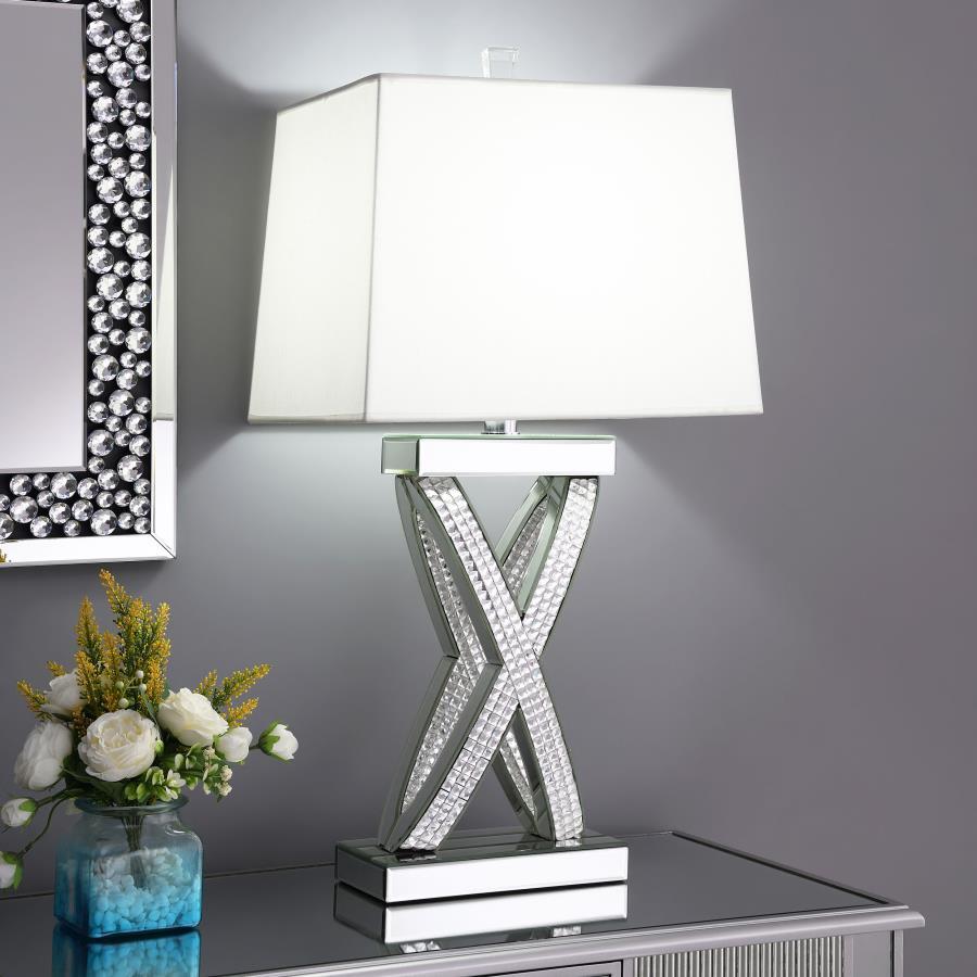 This table lamp features a sculptural X-shaped base accented with shimmering mirrored panels and faceted acrylic crystal inlays. Its bold silhouette and reflective finish create a dramatic presence, while a rectangular white shade provides soft ambient li