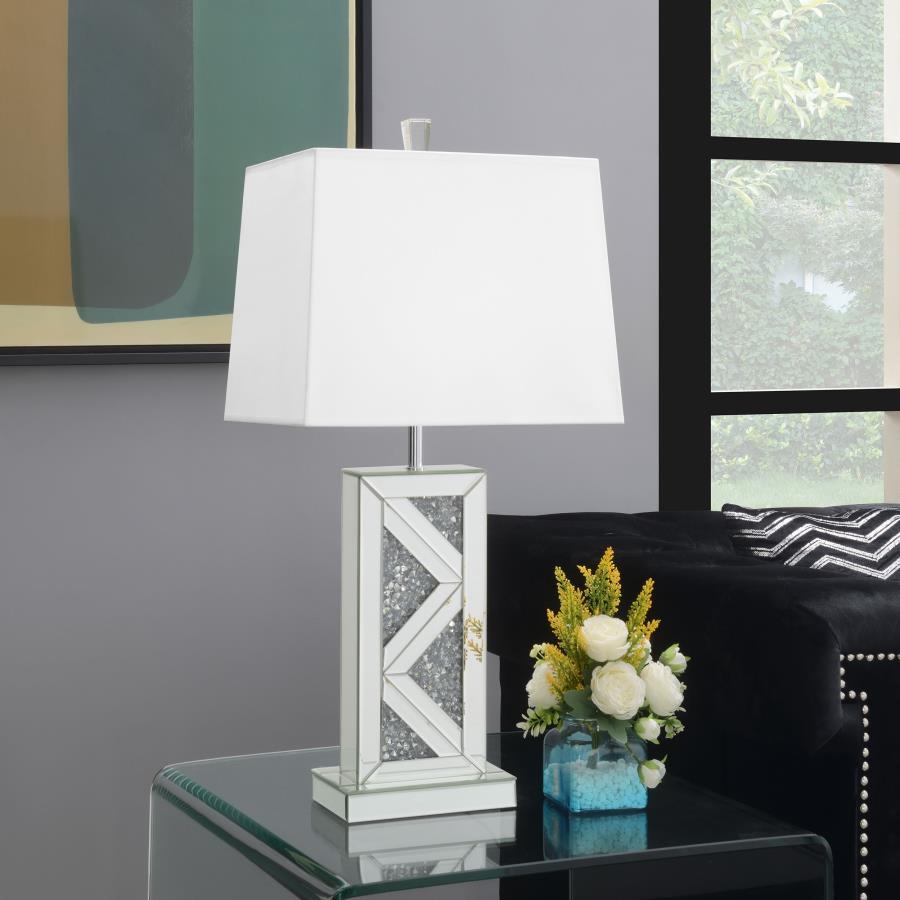 This statement-making table lamp features a rectangular base adorned with mirrored paneling and a bold chevron design inlaid with crushed crystal acrylic. A crisp white shade balances the shimmering base with a clean, structured silhouette. The geometric 